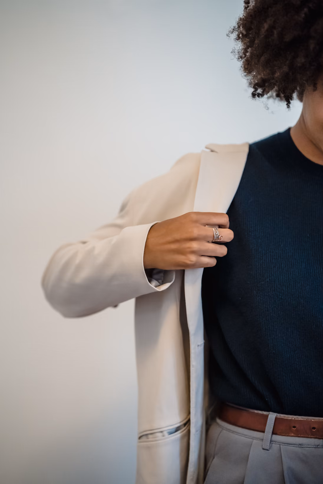 Mulher ajustando um blazer cor de areia sobre uma blusa azul-marinho, com o rosto cortado pela borda do quadro, diante de uma parede branca.